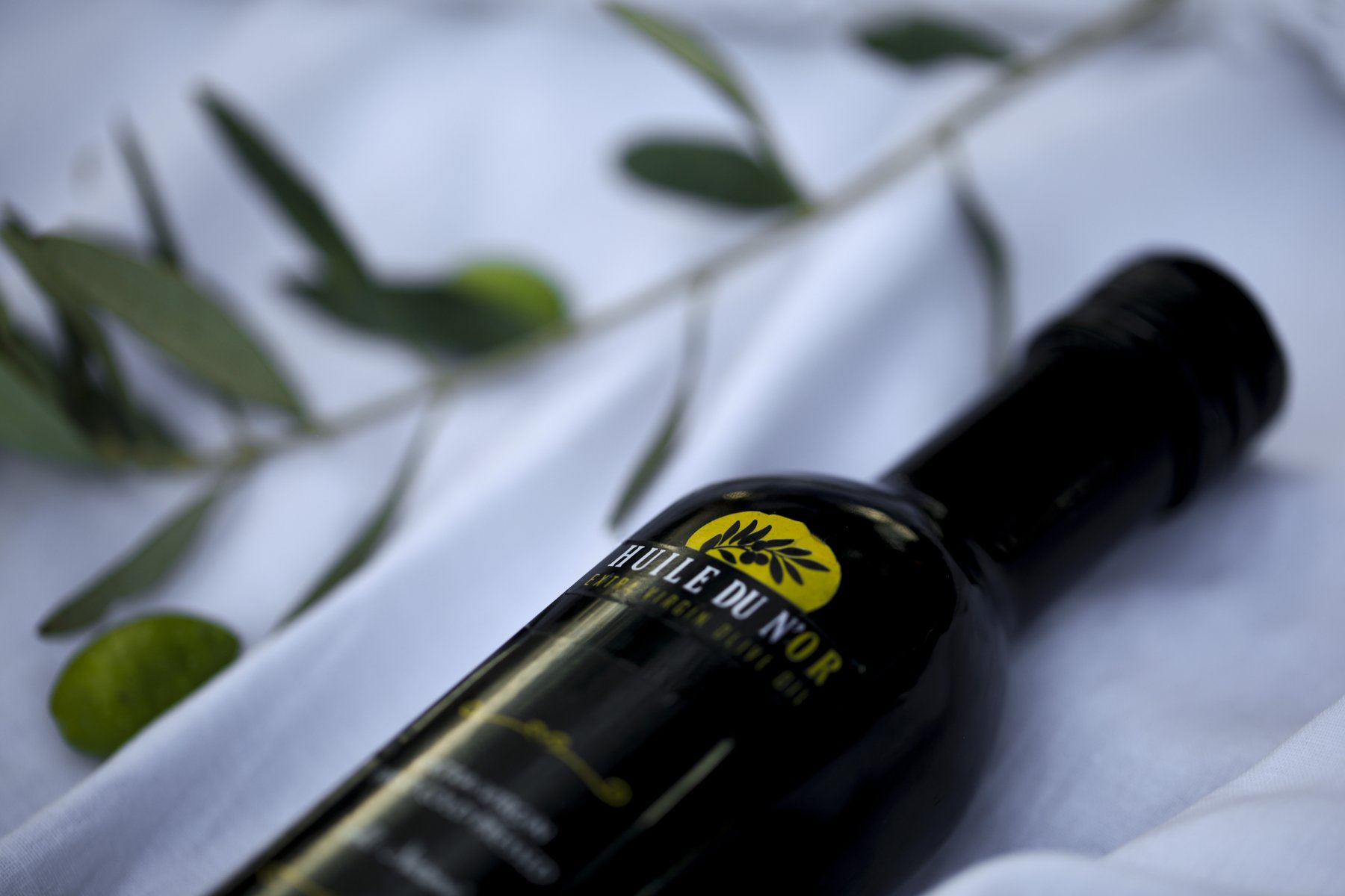 A Huile du N’or bottle on white linen beside an olive branch